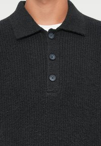 Zwarte, gestructureerde, gebreide polo shirt. Heeft een standaardkraag en drie blauwe knopen. Materiaal lijkt zacht en comfortabel.