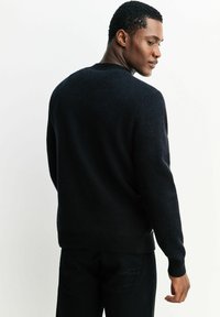 Schwarzer Strickpullover mit rundem Ausschnitt, langen Ärmeln und gerippten Bündchen, der eine strukturierte Oberfläche und minimale Nähte aufweist.