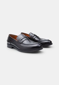 Mocasines de cuero negro con un acabado suave, que presentan acentos de doble correa y un tacón bajo. El interior es marrón con un diseño clásico.