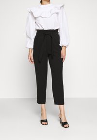 Vit blus med volangkrage och långa ballongärmar, kombinerad med högmidjade svarta byxor och svarta sandaler med öppen tå.