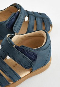 Dunkelblaue Leder-Sandalen mit strukturiertem Stoff, offenem Design, mehreren Riemen und einer braunen Gummisohle. Klettverschlüsse für eine verstellbare Passform.