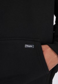 Main dans la poche d'un hoodie noir avec une petite étiquette rectangulaire en argent sur la poche avant.