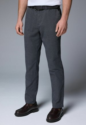 Pantalones grises texturizados con corte recto, que cuentan con un pliegue frontal, bolsillos laterales y un cinturón negro. Combinados con mocasines oscuros.