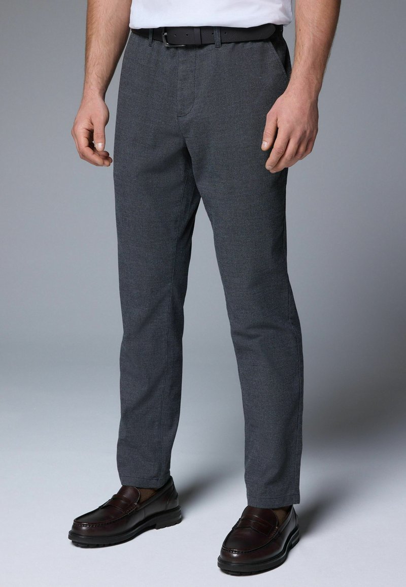 Pantalons gris texturés avec une coupe droite, présentant un pli avant, des poches latérales et une ceinture noire. Associés à des mocassins foncés.