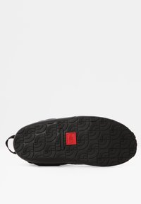 Suela de zapatilla negra con un patrón de agarre texturizado, que presenta un destacado detalle del logo rojo en el centro. Forma redondeada y construcción robusta.