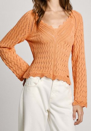 Femme portant un top à manches longues en crochet couleur pêche avec des bords festonnés et un pantalon blanc taille haute, posant avec une main sur la hanche.
