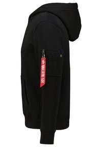 Sweat à capuche noir avec une poche zippée sur la manche et une étiquette en tissu rouge indiquant « Remove Before Flight » attachée à la tirette de la fermeture éclair.