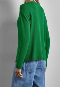 Pull vert en maille avec ourlet côtelé, présentant un col rond et des manches longues, associé à un jean bleu délavé taille haute.