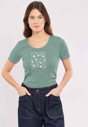 Femme aux longs cheveux bruns portant un t-shirt graphique vert à motifs floraux, rentré dans un pantalon en denim foncé taille haute avec les mains dans les poches.