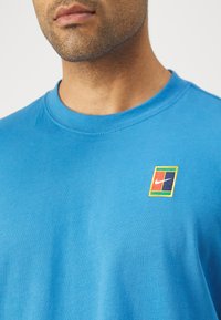Man wearing blue crewneck shirt with small rectangular logo featuring multicolor blocks and white swoosh on chest.