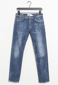 Vaqueros skinny azules desgastados con bolsillos frontales y cierre de botón colgados de una percha negra sobre un fondo blanco.