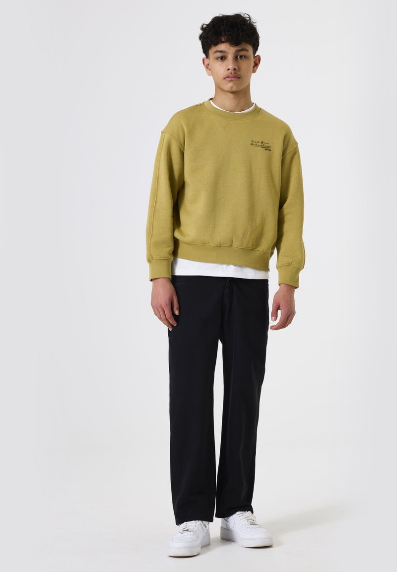 Sudadera de cuello redondo color verde oliva con texto impreso, camiseta blanca debajo, combinada con pantalones rectos negros y zapatillas blancas.