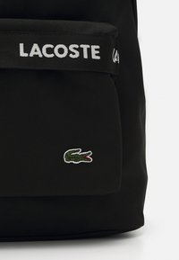 Black fabric backpack with a front pocket, featuring a white "LACOSTE" logo strap and a small green crocodile embroidery detail.