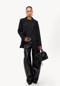 Zwarte gestructureerde blazer over een aansluitend zwart topje, gecombineerd met glanzende zwarte, gemusterde wijde broek en een kleine, gestructureerde handtas.