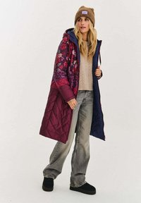 Pikowana kurtka puchowa z kwiatowym wzorem w odcieniach fioletu i bordo, granatowymi rękawami oraz przednim suwakiem. W połączeniu z beżowym swetrem i szarymi jeansami.