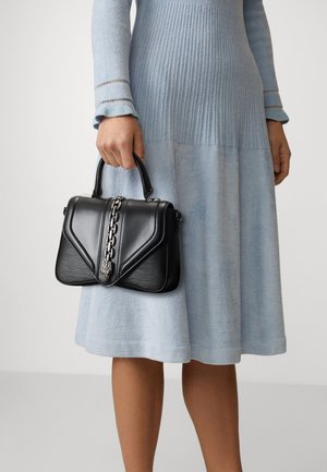 Nainen vaaleansinisessä mekossa, joka pitää kädessään pientä mustaa nahkalaukkua, jossa on ketju ja leijonanpään yksityiskohta.