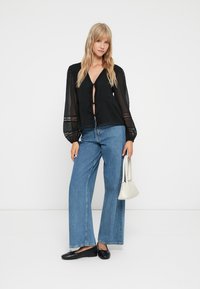 Must mustri blouse läbipaistvate mustriliste varrukatega, seotud ees. Kombineeritud kõrge vöökohaga siniste denimite laiade pükstega ja mustade lamedate kingadega.
