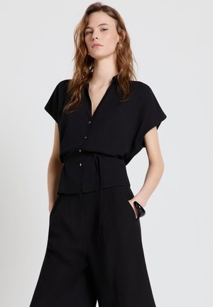 Femme aux cheveux ondulés portant une chemise noire ample boutonnée et un pantalon noir à jambes larges, debout avec une main dans la poche sur un fond neutre.