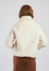 Kremowa kurtka z polaru z miękkim, teksturowanym kołnierzem z futerka. Luźny krój z opuszczonymi ramionami i widocznymi detalami szwów.