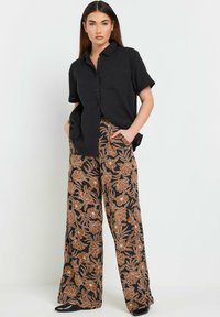 Mustat lyhythihaiset nappipaidat yhdistettynä leveisiin kukkakuosisiiin housuihin, joissa on ruskeaa ja mustaa väriä. Malli seisoo kädet taskuissa.