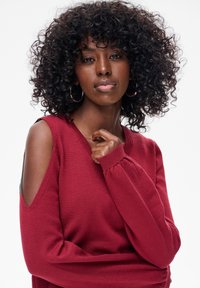 Kobieta o kręconych włosach i kolczykach-kołach, ubrana w czerwony sweter z odkrytymi ramionami, patrząca pewnie w obiektyw.