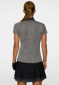 Vrouw met een paardenstaart, gekleed in een aansluitend grijs shirt met korte mouwen en een zwarte plissérok, gezien vanuit de rug tegen een effen achtergrond.