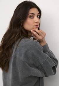 Pilkas didelis džemperis su tekstūruotu audiniu, turintis apvalią iškirptę, raišteliais aptrauktus rankogalį ir laisvą kirpimą, rodomas šonu.