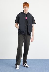 Μαύρο γουλιέ βεστ με ψηλό γιακά, φορεμένο πάνω από ένα μαύρο t-shirt με πολύχρωμο λογότυπο, συνδυ�ασμένο με σκούρους παντελόνες και μαύρα-άσπρα αθλητικά παπούτσια.