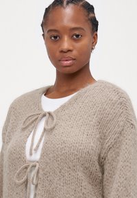 Beige neule Cardigan, jossa on teksturoitu kuvio, avonainen etuosa ja kaksi nauhakiinnitystä kaulan kohdalla. Pehmeä, pörröinen materiaali ja väljä malli.