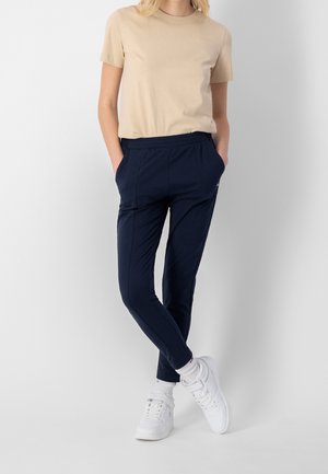 Person med beige kortærmet skjorte, navy slim-fit bukser, hvide sokker og hvide sneakers, stående med hænderne i lommerne mod en hvid baggrund.