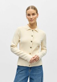 Femme portant un cardigan crème boutonné avec des boutons dorés et un jean bleu, debout devant un fond clair uni.