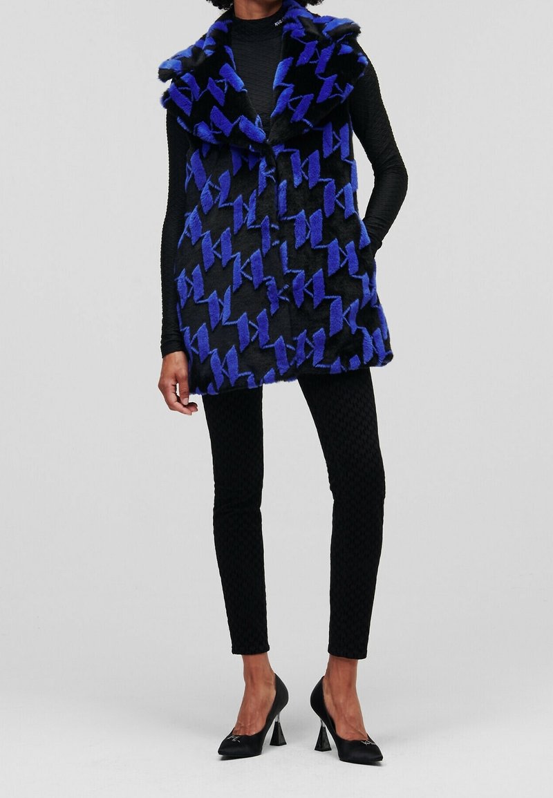 Femme portant un gilet sans manches en fausse fourrure à motif noir et bleu, par-dessus un haut noir à manches longues, un pantalon noir texturé et des escarpins noirs.