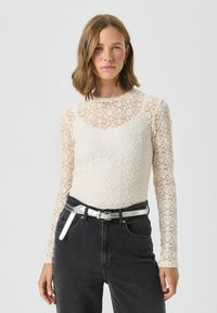 Mežģīņu garām piedurknēm augšdaļa off-white krāsā ar ziedu rakstu; noapaļota kakla izgriezums; apvienota ar augstas jostasvietas melnajām džinsām un sudraba jostu.
