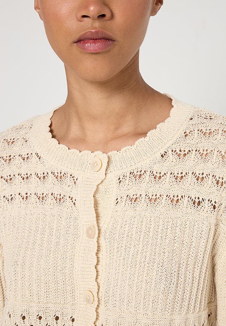 Dzianinowy kremowy kardigan z falbaniastym dekoltem, z zapięciem na guziki, koronkowymi wzorami i teksturowanym ściągaczem.
