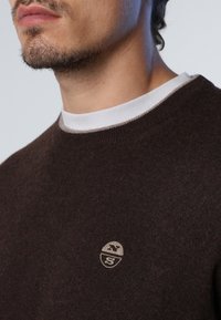 Pull marron tricoté avec un col rond, présentant un col intérieur blanc et un petit logo sur la poitrine. Texture douce et coupe classique.