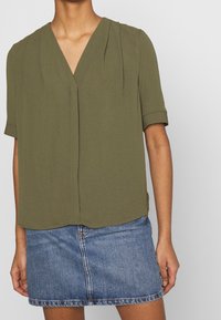 Donna che indossa una blusa a maniche corte verde oliva con plissettature e scollatura a V, abbinata a una minigonna in denim blu.