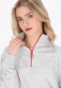 Šedá mikina na zip s vysokým límcem a červeným zipem, vyrobená z texturované tkaniny a s dlouhými rukávy.