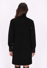 Schwarzes gestricktes Kleid mit hohem Kragen, gerippter Textur oben und verzierten Ärmeln, die farbige Nieten aufweisen.
