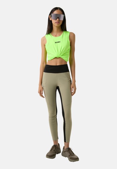 Neon green cropped tank top with knot detail, paired with beige leggings featuring black side panels. Model wears oversized reflective sunglasses and gray sneakers.