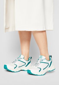 Chaussures de sport blanches avec des accents turquoise, tige en maille texturée et col rembourré. Elles se caractérisent par une semelle épaisse et rembourrée et un système de laçage.