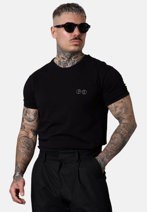 Mann mit Tätowierungen an Armen und Hals, trägt schwarze Sonnenbrille, schwarzes eng anliegendes T-Shirt und schwarze Hose, posiert mit einer Hand in der Tasche.