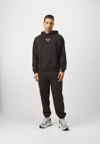 Hanorac negru și pantaloni joggers asortați, dintr-un amestec de bumbac. Hanoracul are o buzunar frontal și detalii metalice cu logo. Ghete sunt gri și albe.