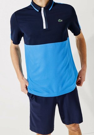 Mann trägt ein marineblaues und hellblaues kurzärmeliges Poloshirt mit Knopfleiste und marineblaue Shorts, steht mit den Händen in der Nähe der Taschen.