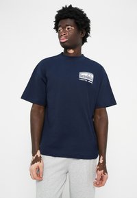 Mørkeblå bomuld t-shirt med korte ærmer, med et logo-patch foran. Bæret sammen med grå sweatpants, der har en afslappet pasform.