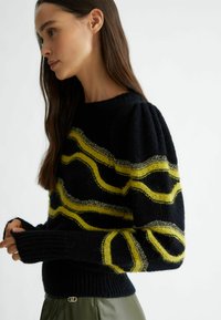Maglione nero a maglia con strisce ondulate gialle e dettagli argentati. Le maniche a sbuffo e i polsini a coste arricchiscono la texture e il design.