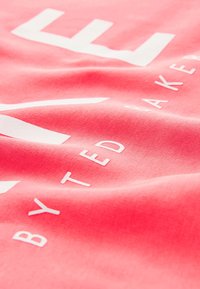 Camiseta de algodón color coral con una textura suave, que presenta grandes letras blancas que dicen "LIFE" y "BY TED BAKER" en un diseño minimalista.