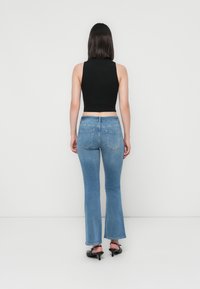 Γυναίκα με καστανά μαλλιά μέχρι τον ώμο, φορώντας ένα αμάνικο μαύρο crop top, ανοιχτό μπλε φαρδιά τζιν και μαύρα τακούνια, στέκεται μπροστά σε έναν λευκό τοίχο.