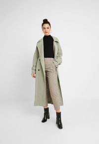 Long olive green trench coat over a black turtleneck, paired with beige and brown checked high-waisted trousers and black heeled boots.