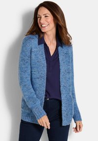 Frau lächelt, trägt eine blaue Strickjacke über einem dunkelblauen Hemd mit Kragen und dunklen Hosen, steht vor einem hellen Hintergrund.
