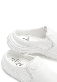 White slip-on shoes made of smooth synthetic material, featuring elastic side panels, a rounded shape, and a textured rubber sole.
